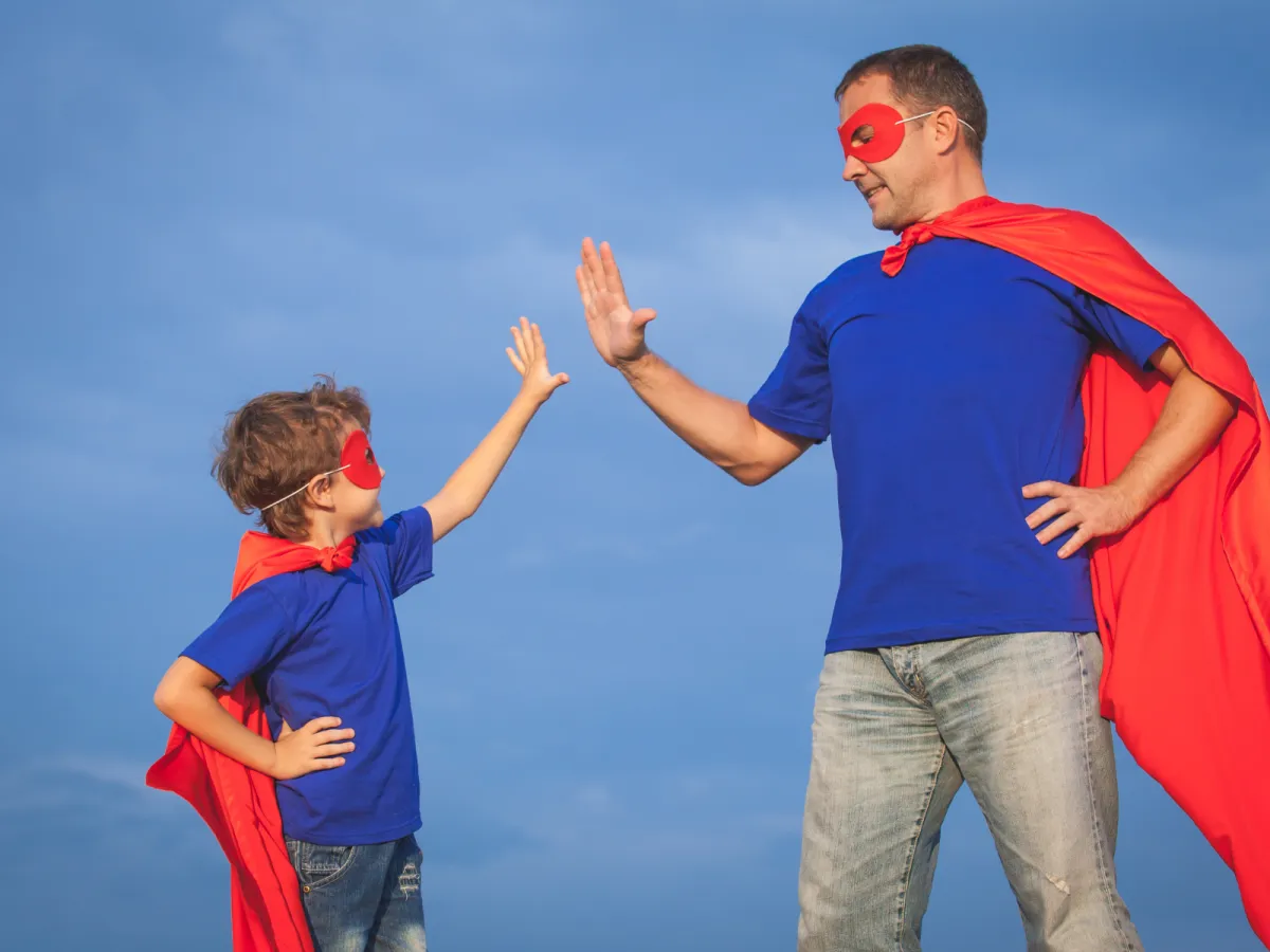 Adult and child wearing superhero capes and masks, smiling and high-fiving outdoors under a blue sky, symbolising confidence and connection