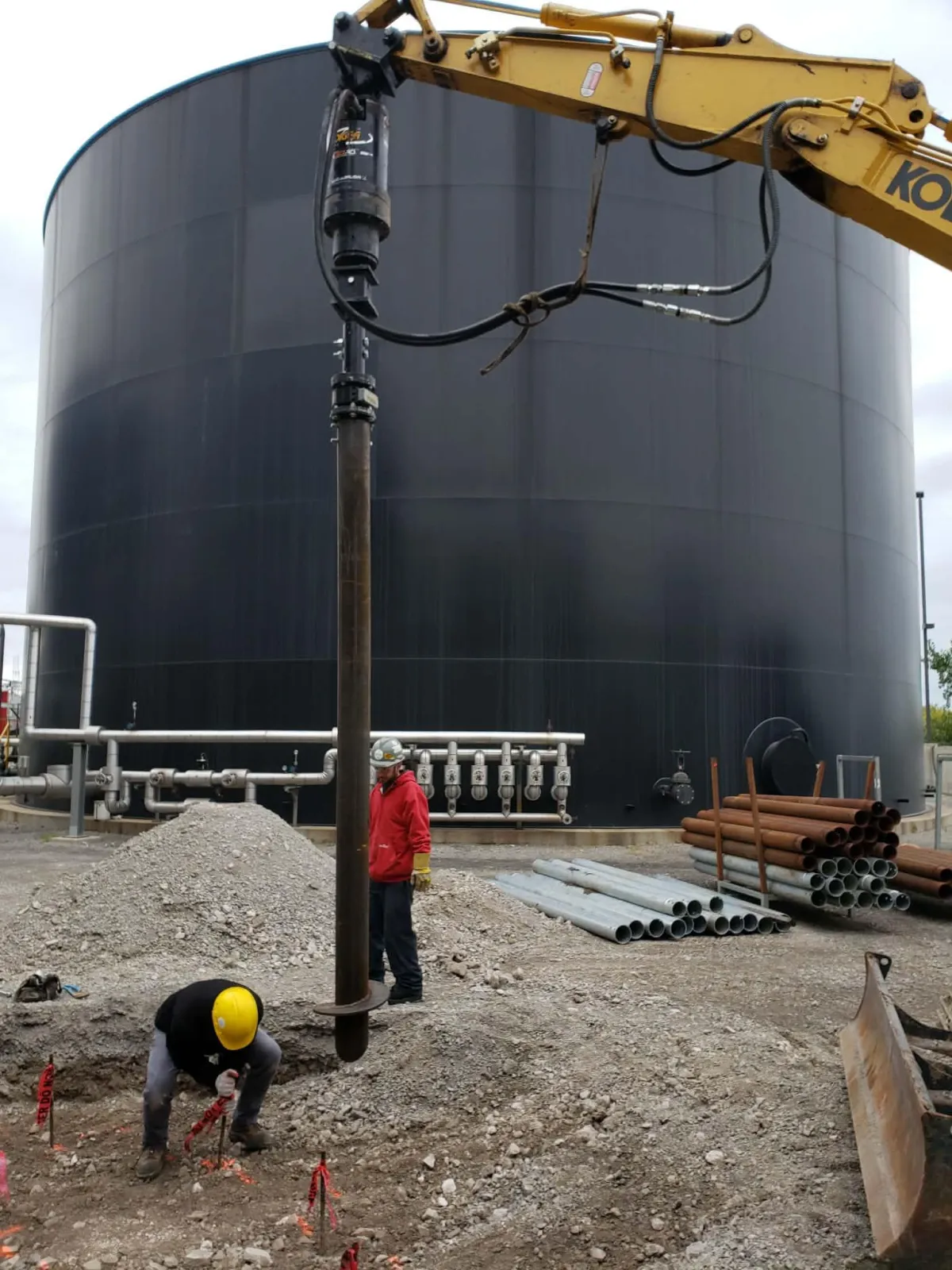 Crew installing a large industrial screw pile foundation beside chemical storage tanks at a commercial job site.