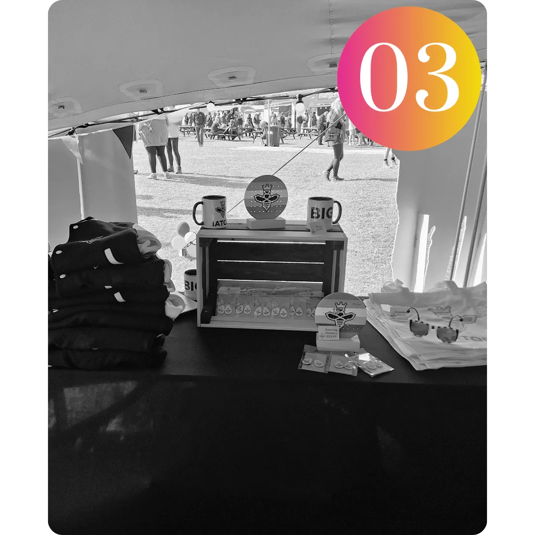 Image of the IATQB set up at Ideas Fest - showing Varsity Jackets folded, mugs, earrings and an earring holder with a pair of bee sunglasses on tote bags