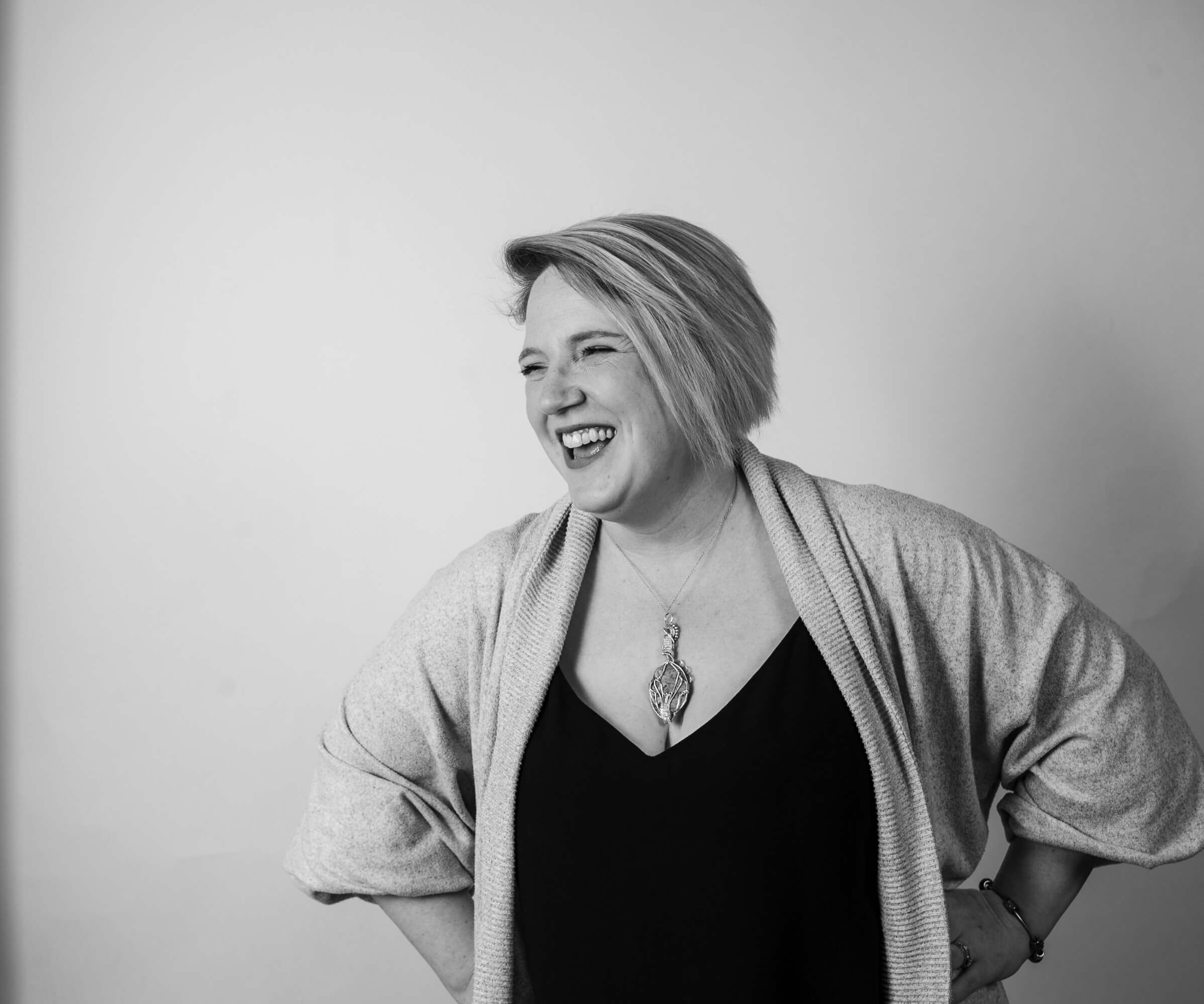 Photo of Lisa Dalgleish - a white woman with short hair wearing a black top and grey cardigan - standing against a wall smiling. Photo is in greycale