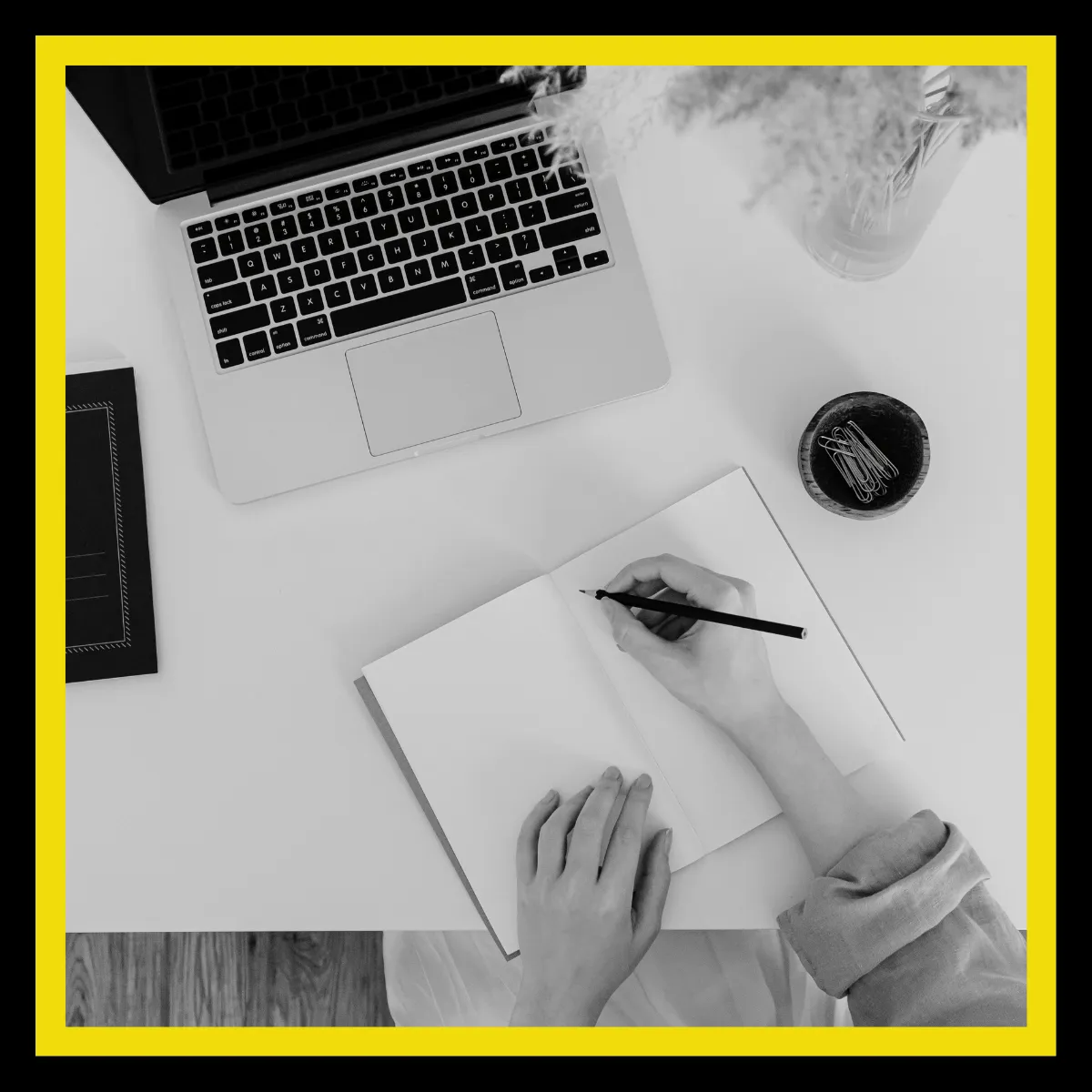 Picture of two hands on a notebook at a desk writing with a laptop at the of the image (greyscale)