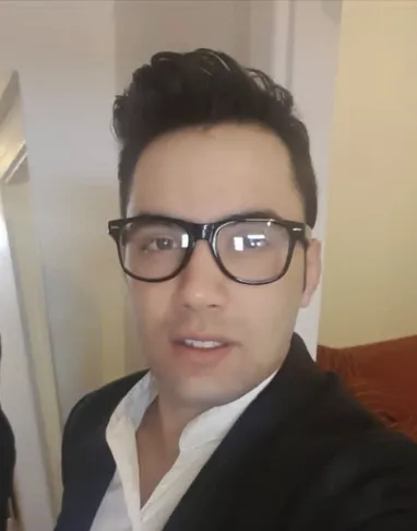 A middle-aged man with glasses and a gold tie, seated at a desk with property documents, looks directly at the camera, smiling. The background is a bright office with black and gold decor, suggesting professionalism and trust.