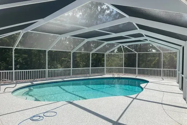 Screened-in backyard pool enclosure with a white aluminum frame, turquoise swimming pool, and surrounding trees.