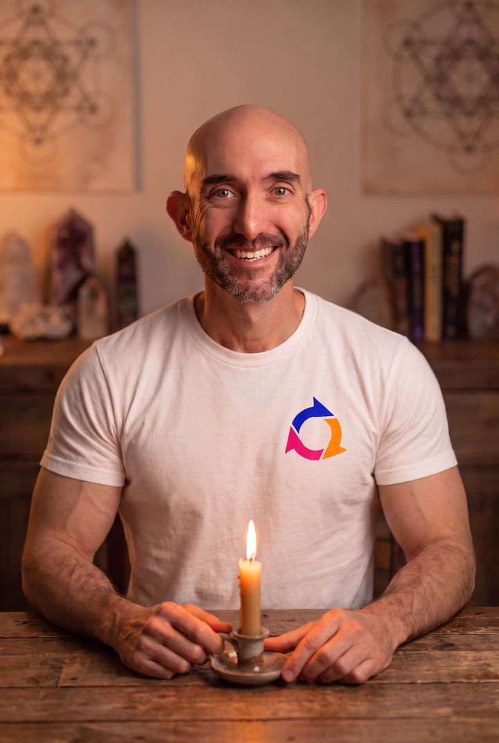 A handsome Mojoe Mike sits at a wooden table. We see him from the waist up. On the table is a lit stick candle that Mike is holding. He is smiling at the camera and wearing a white Spin Positive Tri-Arrow logo t-shirt. Behind him are various books and tapestries along the wall.