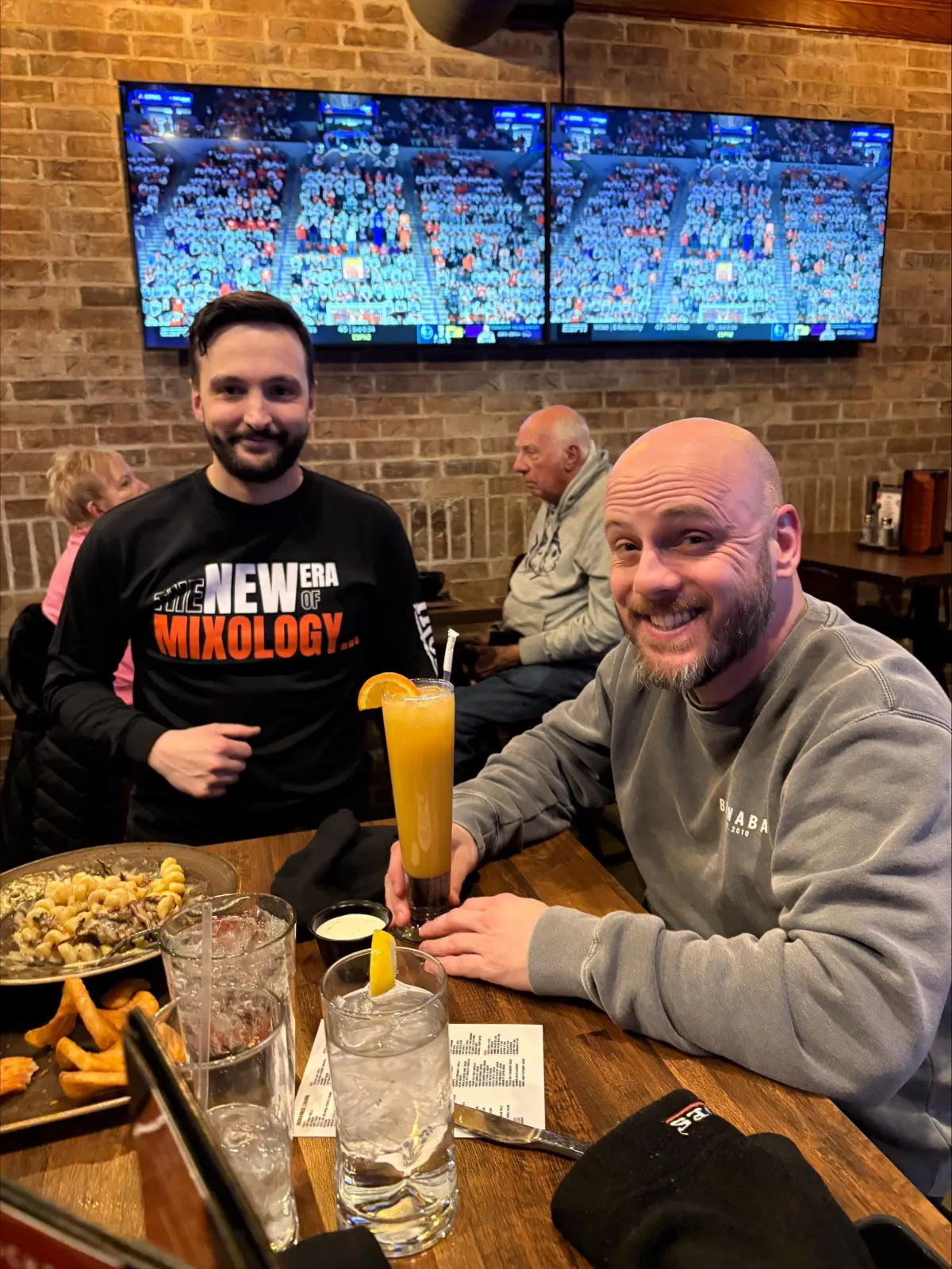 Two men enjoying food and non-alcoholic cocktails at a bar while watching a sports game. One man wears a shirt reading “The New Era of Mixology,” reinforcing the Sober Sips tagline, “Mood-Infused Cocktails. The Future of Social Drinking.