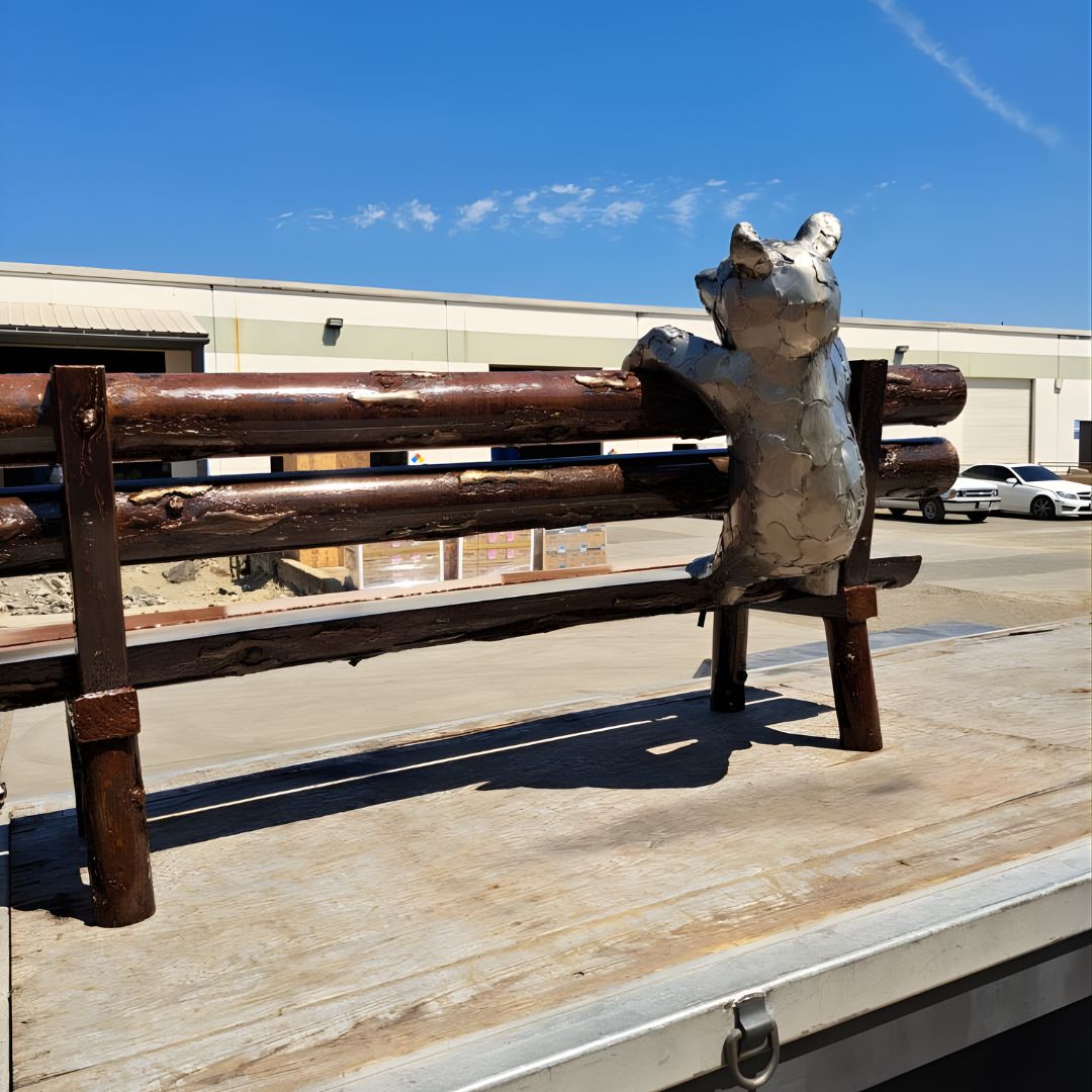 A side view of a similar dark-stained wood bench sitting on a flatbed trailer. A metal bear sculpture is attached to the back, appearing to peek over the top rail.