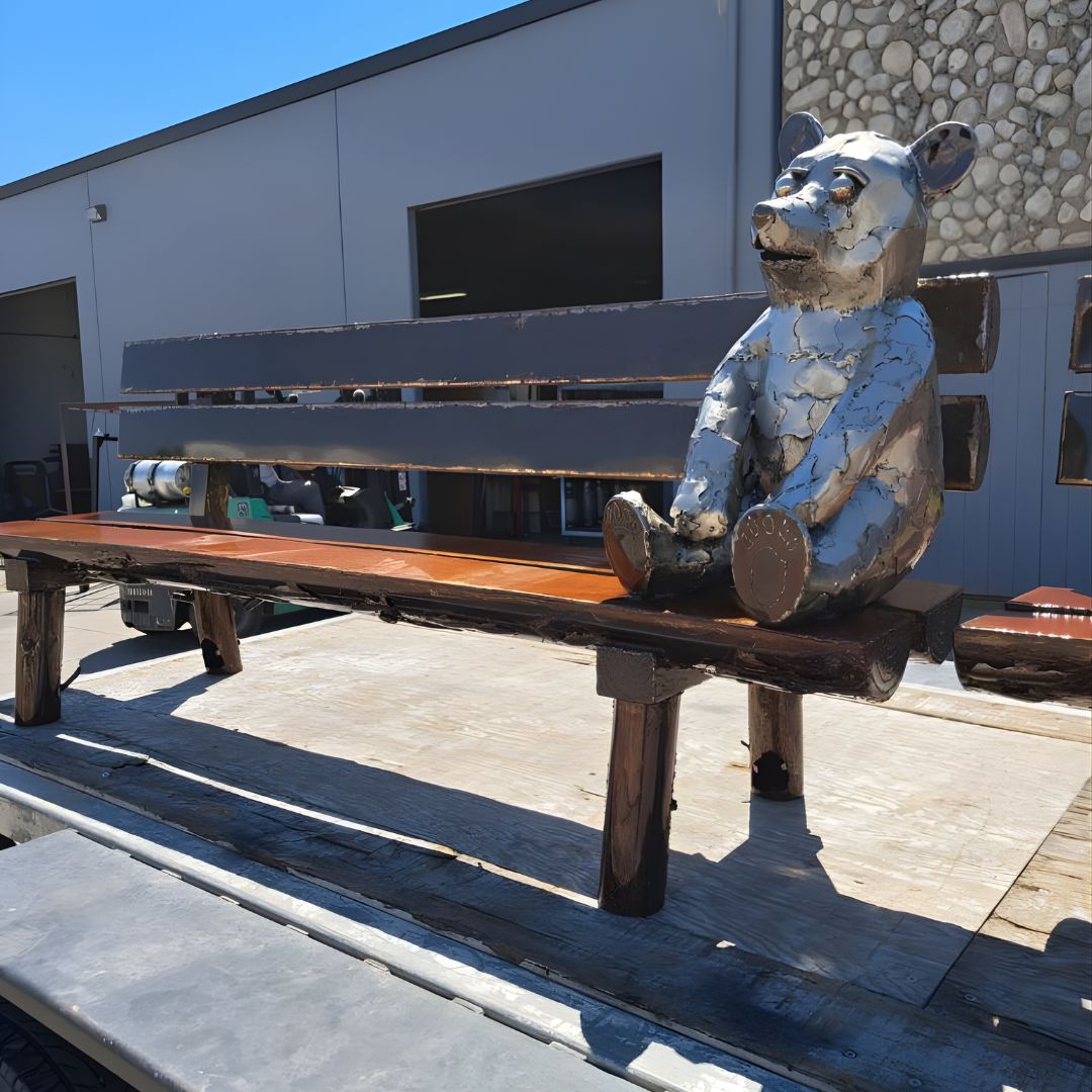 A full view of a completed outdoor bench featuring dark wood planks and a metallic bear sculpture sitting upright on one end of the seat.