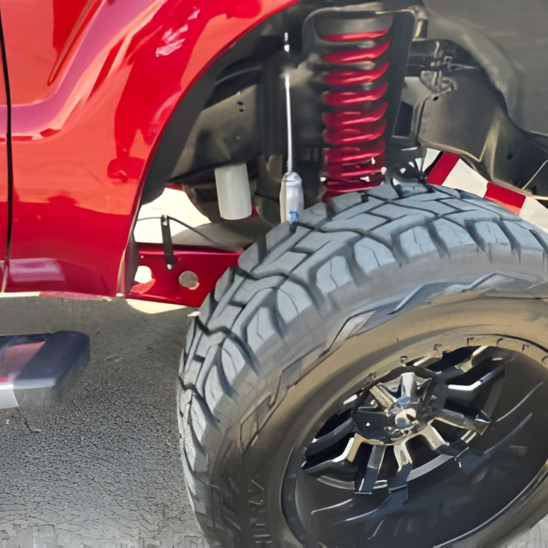 A close-up underneath a lifted red truck, showing an aggressive off-road tire on a black rim, along with a bright red coil spring and upgraded suspension shocks.