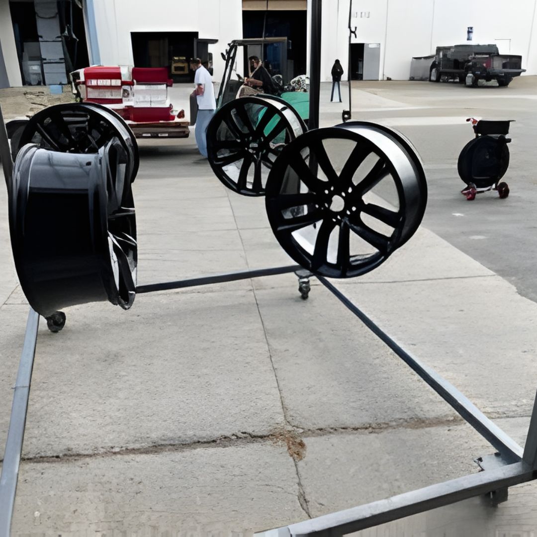 Four large, black custom car rims hanging on a rolling metal rack, with a shop exterior in the background