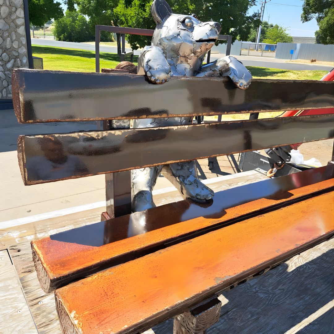 A close-up of a rustic wooden bench with a dark, glossy wood finish. A metallic silver bear sculpture is designed to look as though it is climbing over the backrest.
