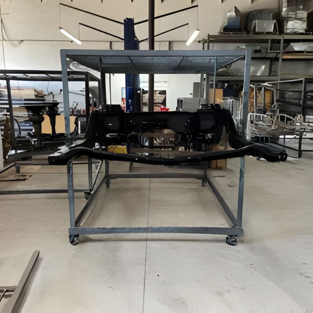A bare vehicle chassis painted black, sitting on a large rolling cart inside a metal fabrication or paint shop.