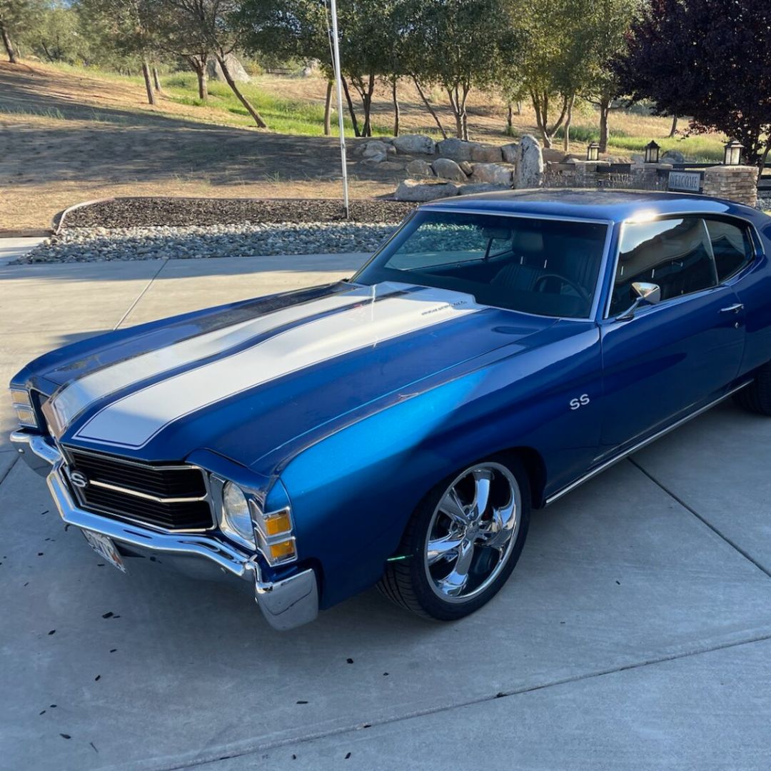 A classic blue muscle car (resembling a Chevrolet Chevelle) featuring dual white racing stripes down the hood, parked outdoors on a concrete driveway.