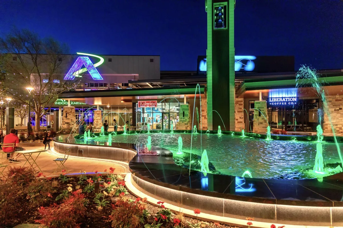 Vibrant nighttime scene at a Frisco, Texas plaza featuring a colorful fountain, illuminated buildings, and people enjoying the outdoor space, highlighting local businesses like Liberation Coffee Co. and Kilwins.