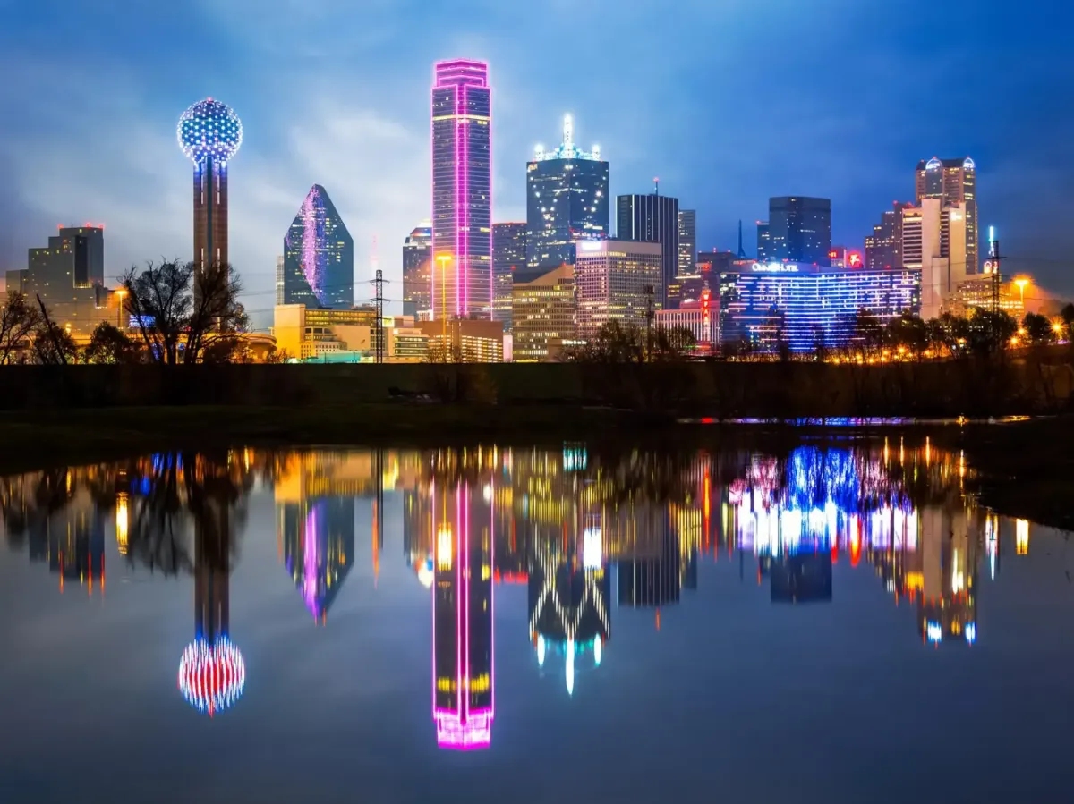 Dallas skyline at night with colorful lights reflecting in water, featuring notable landmarks like the Reunion Tower and modern skyscrapers, relevant to OttoMate 360's services in the Dallas-Fort Worth area.