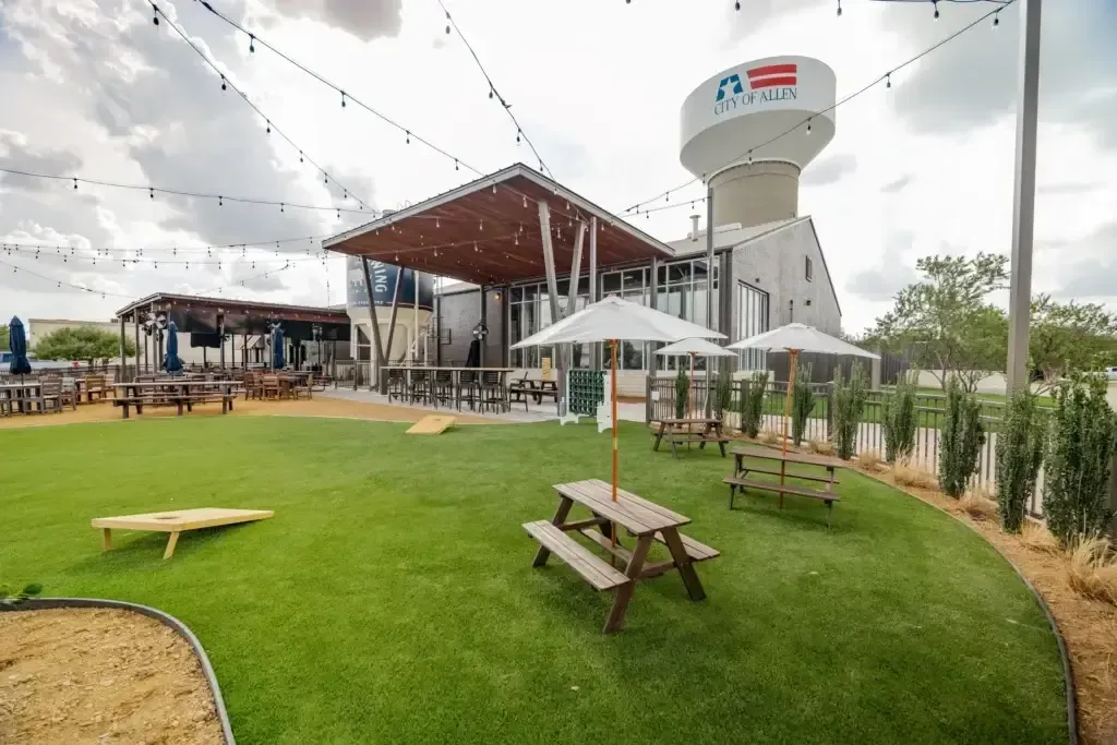 Outdoor seating area at a venue in Allen, Texas, featuring picnic tables, umbrellas, and a visible water tower labeled "City of Allen," surrounded by green grass and string lights.