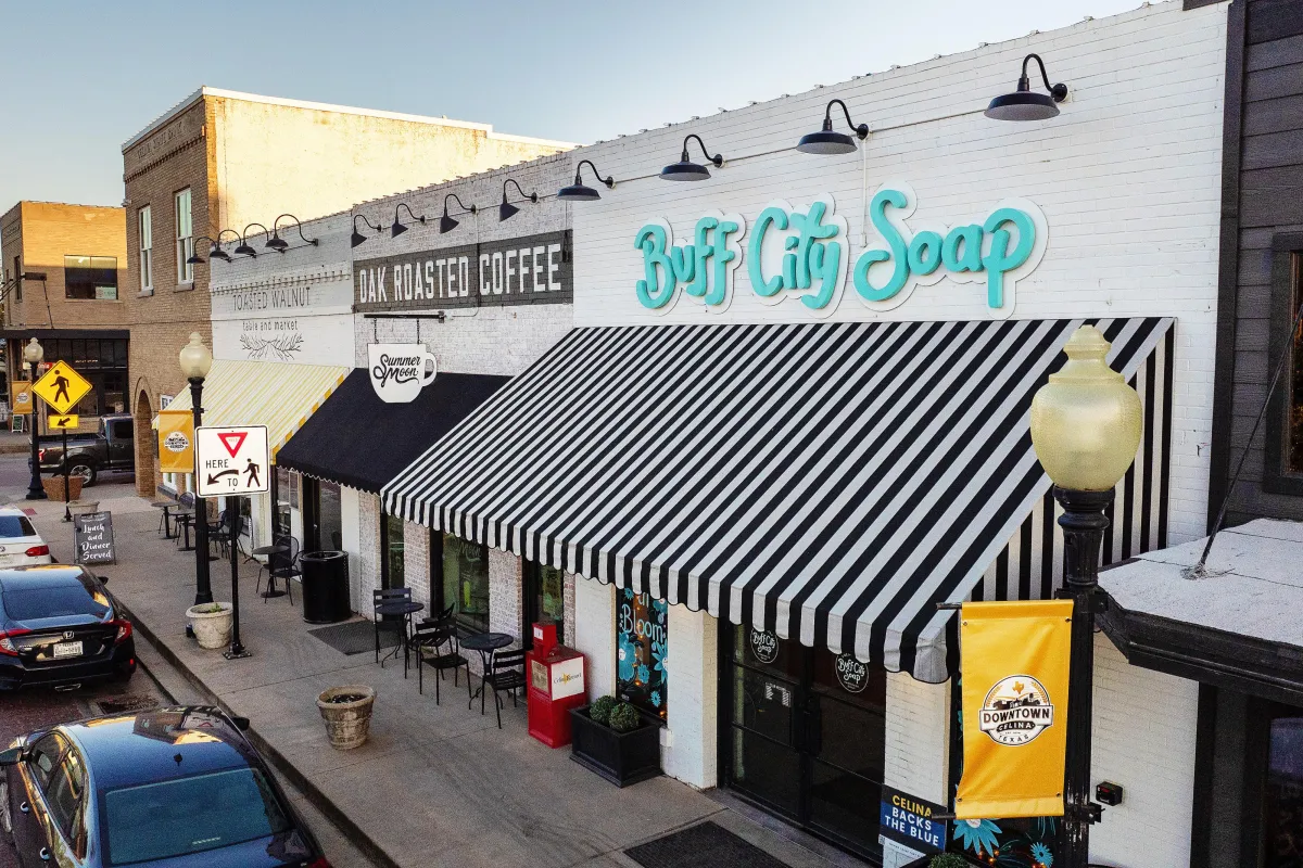 Downtown storefronts in Celina, Texas featuring Buff City Soap and Oak Roasted Coffee, with striped awnings and outdoor seating, highlighting local business atmosphere.