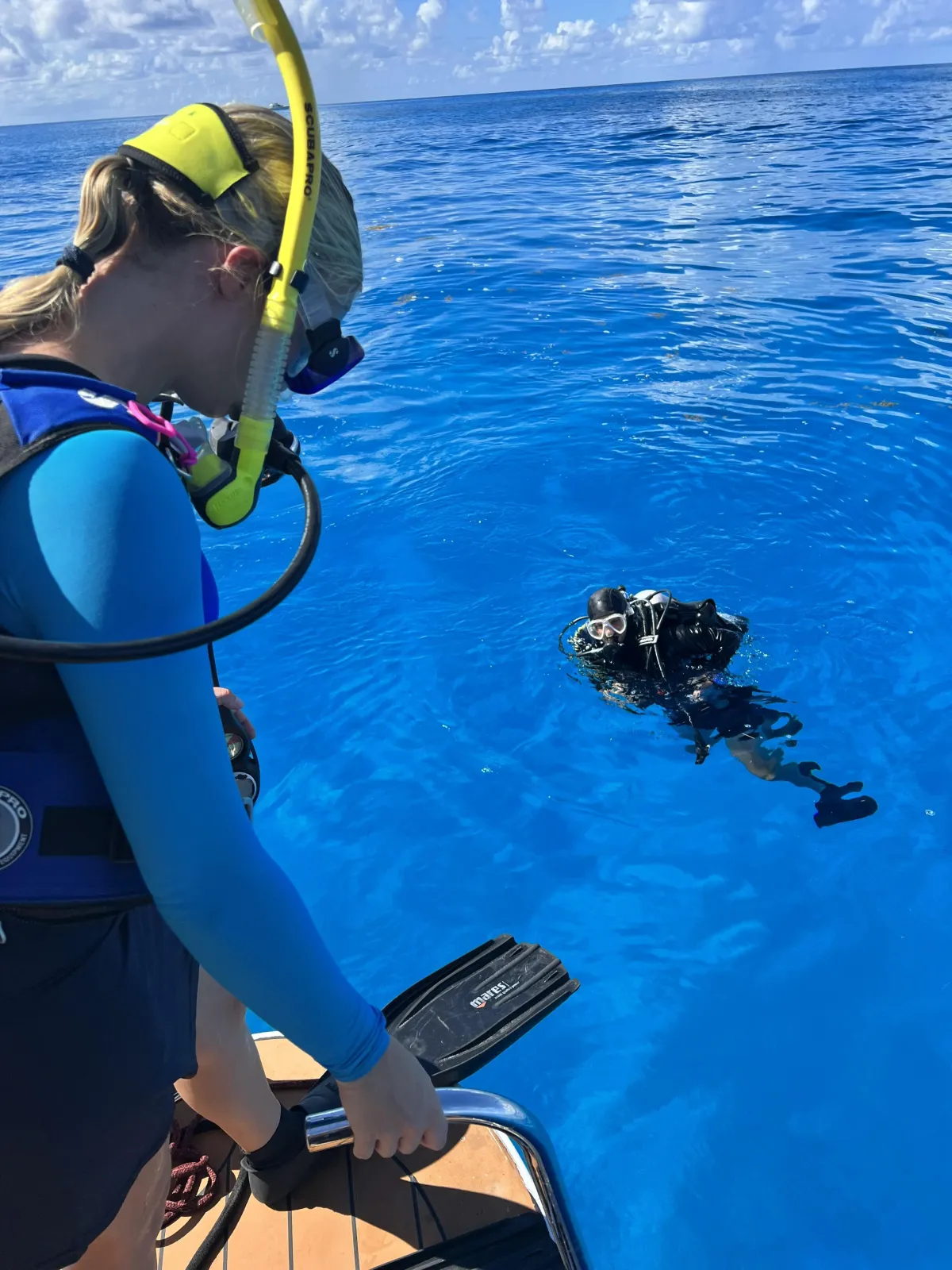 A diverse group of students and an instructor gathered poolside, reviewing dive equipment and safety procedures. The setting is bright, with clear blue water and educational materials visible. 3:2 aspect ratio.