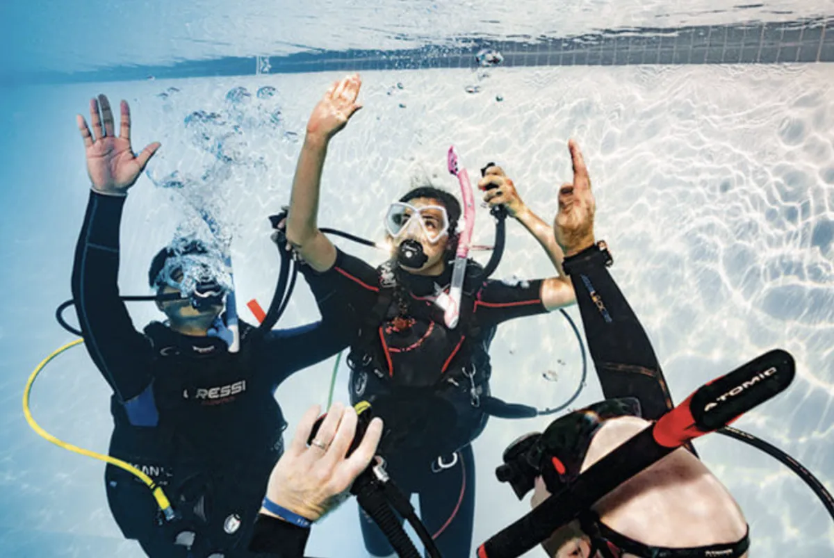 A seasoned dive instructor, female, mid-40s, demonstrating rescue techniques to a diverse group of adult trainees on a sunlit pool deck. The group is attentive, wearing wetsuits, with certification manuals visible, set against a backdrop of palm trees and blue sky.