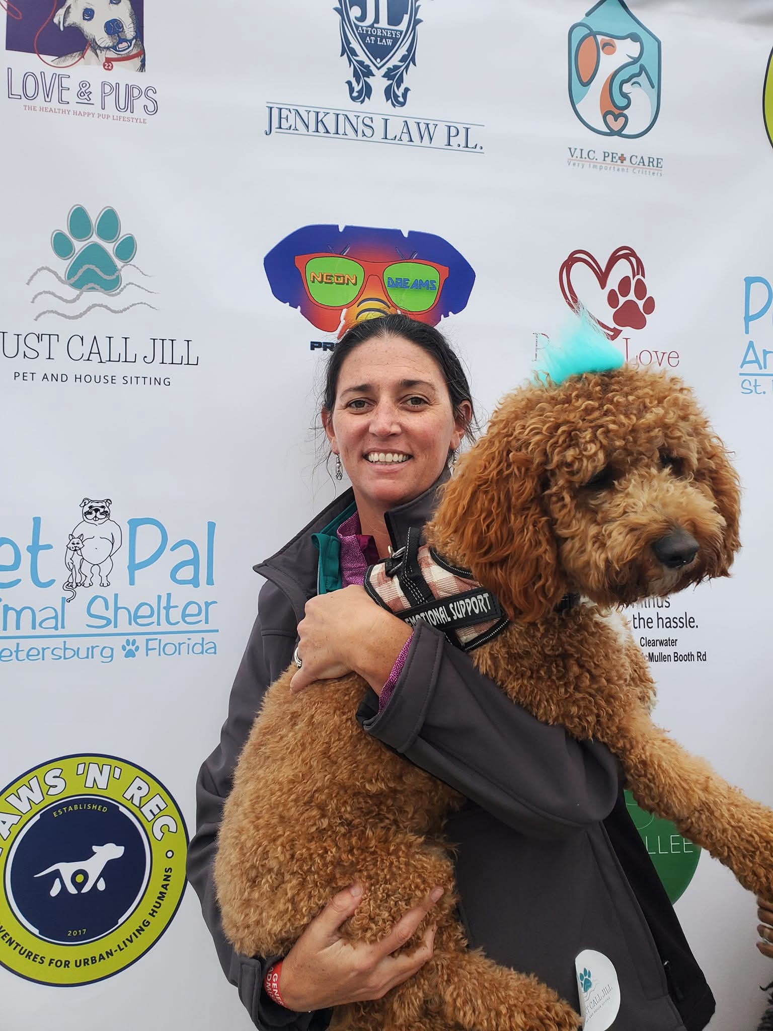 Woman holding a curly brown service dog in front of pet event sponsor backdrop.