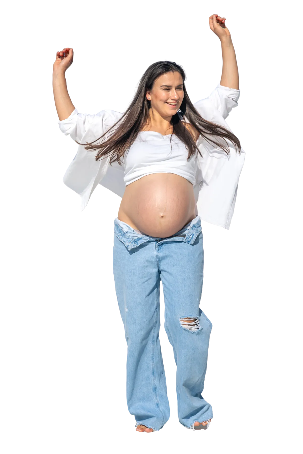 Pregnant woman joyfully dances, arms raised, wearing a white top and unbuttoned jeans. Her expression is happy and carefree, set against a plain background.
