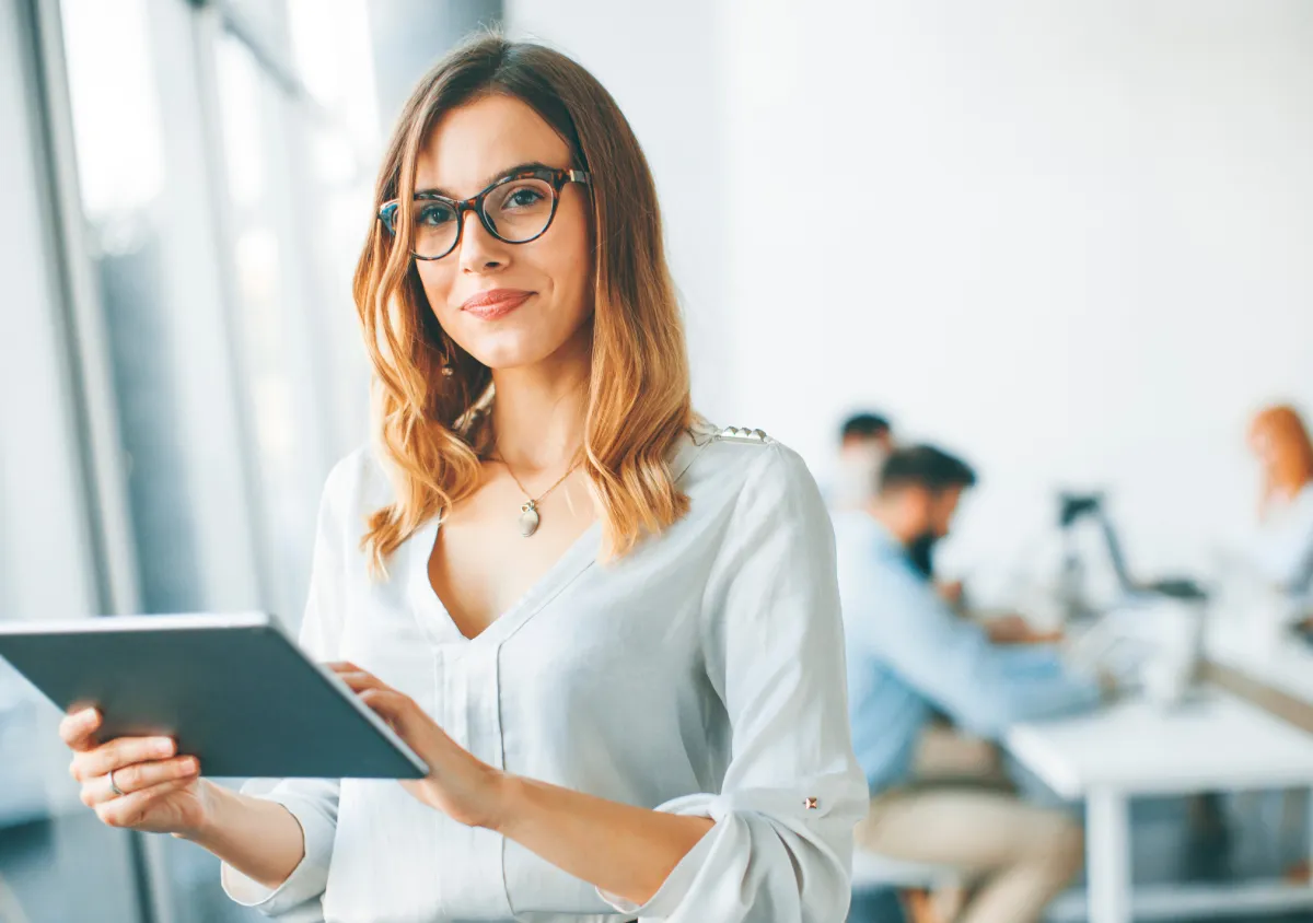 Woman with a tablet looking at the camera with a work space behind her