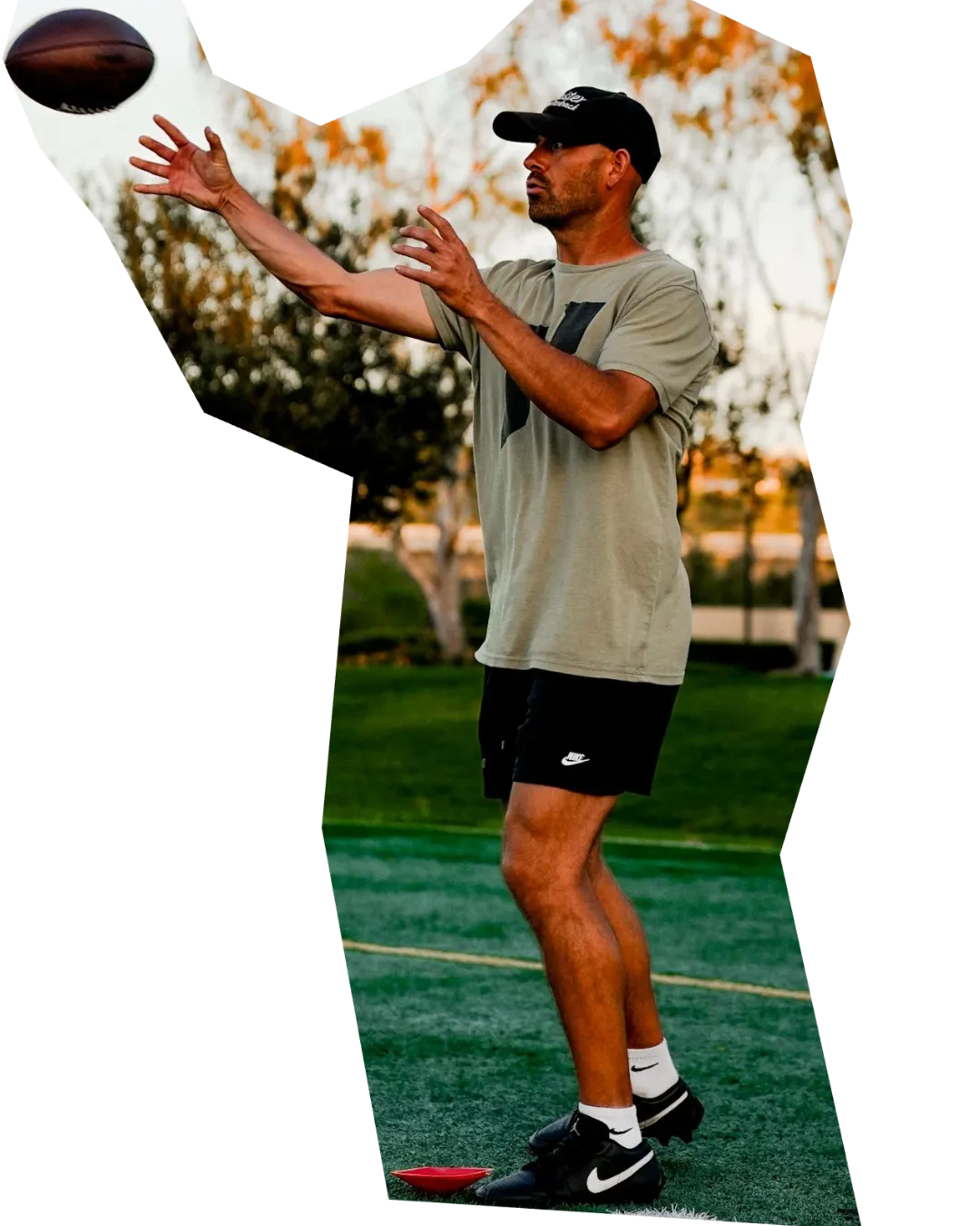 A man wearing a tan t-shirt, black shorts, a black baseball cap, white socks, and black sneakers is standing on a grassy field, reaching up with both hands to catch a dark brown football that is suspended in the air above his right hand. His body is slightly turned to his left. Two small, colorful objects, possibly cones, one red and one yellow, are on the ground near his feet. The background shows a park setting with trees and foliage, some of which have autumn colors, under a bright sky.