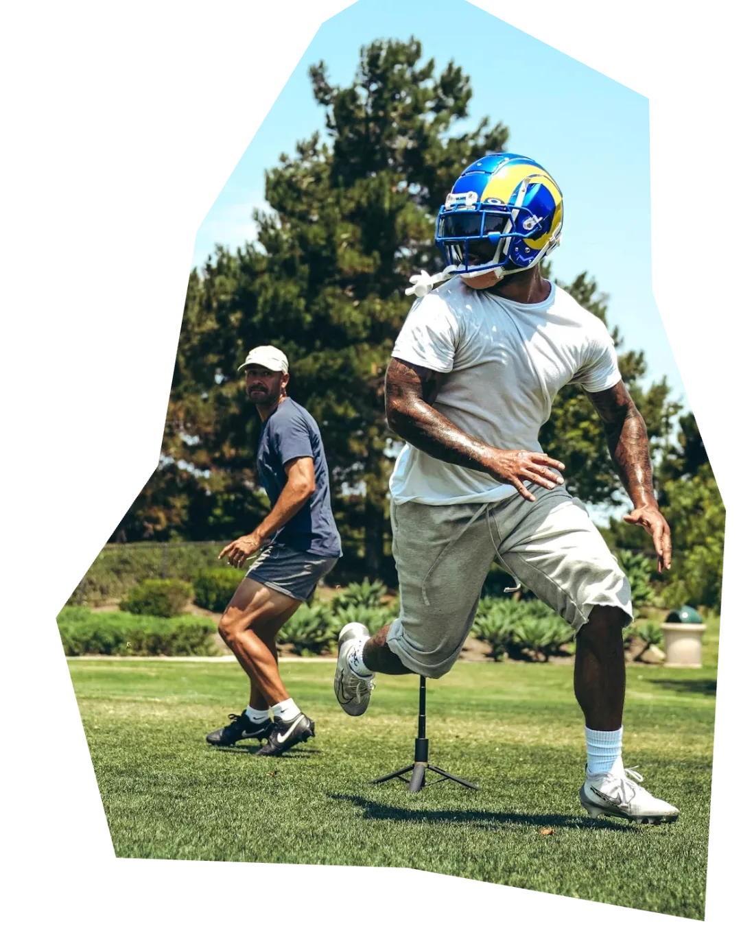 A football player wearing a Los Angeles Rams helmet, a white t-shirt, and light gray shorts is running on a grassy field. The player is also wearing white socks and white cleats. His right arm has visible tattoos. To his left and slightly behind him is an instructor wearing a dark blue shirt, dark shorts, a white baseball cap, and black sneakers, who appears to be watching or following the player. A small, black agility pole is visible between the player's legs. The background features tall trees and bushes under a bright blue sky.