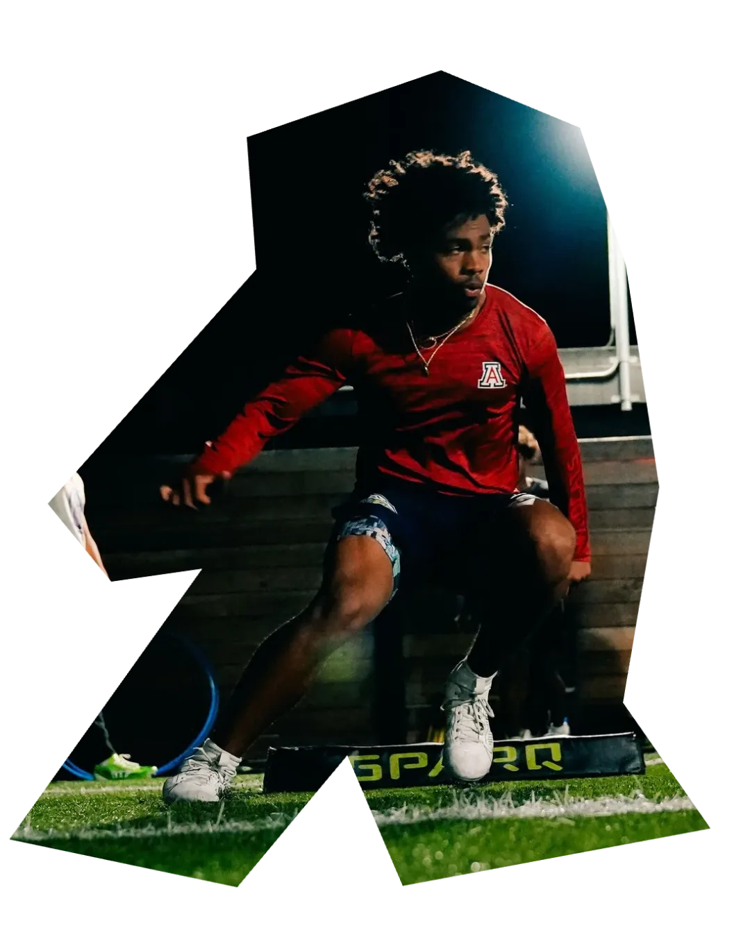 A football player from WR1 Performance with dark, curly hair wearing a red long-sleeve shirt and dark shorts is training at night on a turf field under a bright field light. He is positioned low to the ground with his weight shifted forward, suggesting a drill focused on agility or explosion. The word "SPARQ" is visible on a piece of training equipment he is stepping over. The image also contains the acronym "LFA" and other individuals walking in the background.