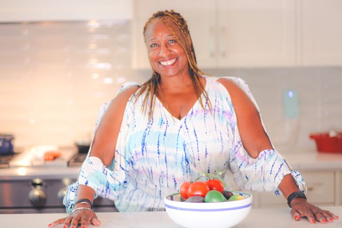 Smiling woman with braids in a bright kitchen holding a bowl of fresh produce.