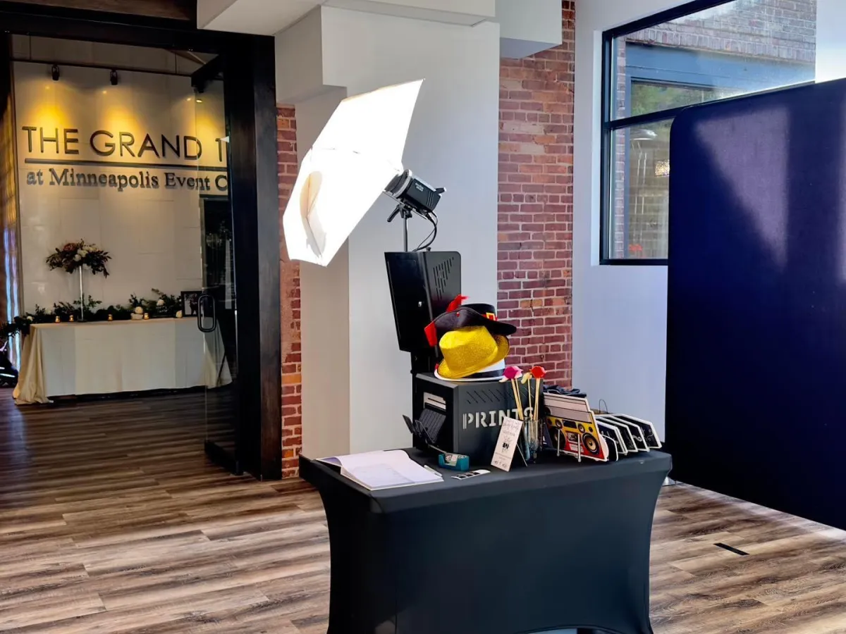 A festive photo booth setup with gold and red decorations, a group of diverse professionals in business attire laughing and posing with holiday props, set in a modern office event space. Warm lighting and branded signage complete the scene.