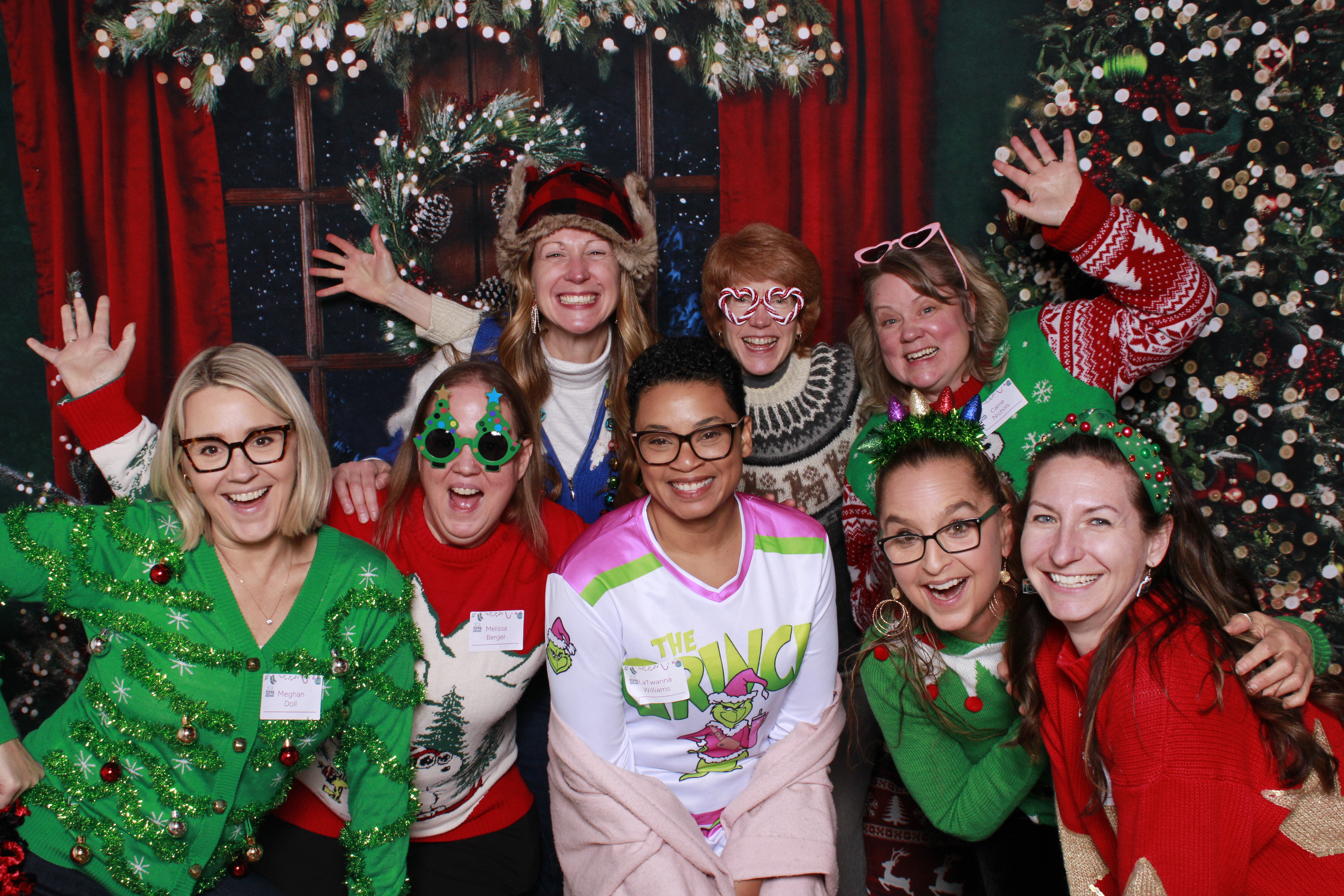 A candid moment of employees in festive sweaters using a photo booth, holding branded props and laughing, with a decorated office background and twinkling string lights. The group is diverse in age and ethnicity.