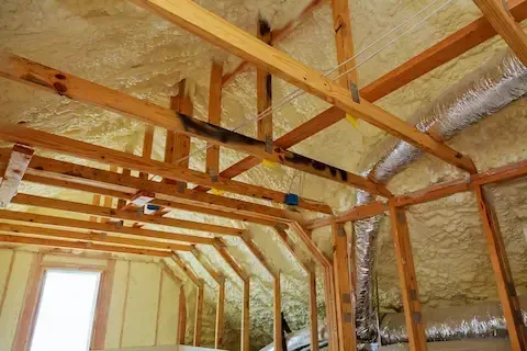 Attic rafters and roof deck coated in open-cell spray foam with silver ductwork running below