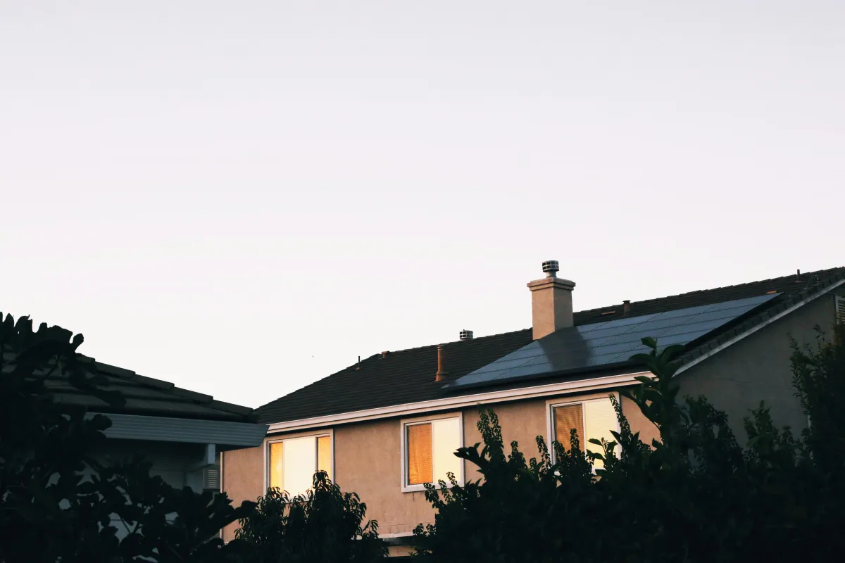 A modern house with solar panels installed on the roof, captured in soft, cool evening light, with surrounding trees partially framing the image.