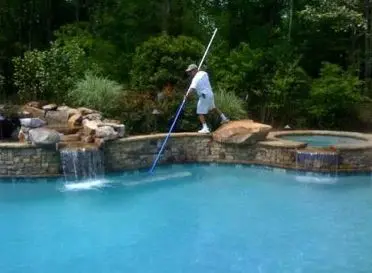 Technician vacuuming pool floor with telescoping pole beside stone waterfall and raised spa