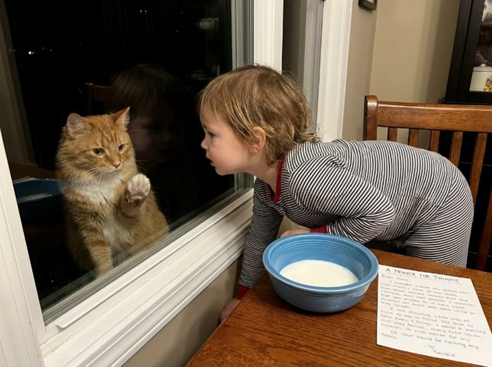 Little girl looking through a window at her orange cat beside a bowl of milk and a printed poem