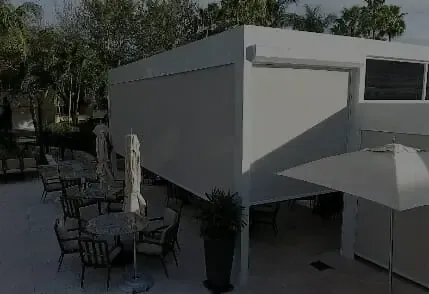 An outdoor patio area with tables and chairs under opened umbrellas beside a modern white architectural structure with large windows and landscaped bushes, equipped with retractable bug screens to protect your guests.