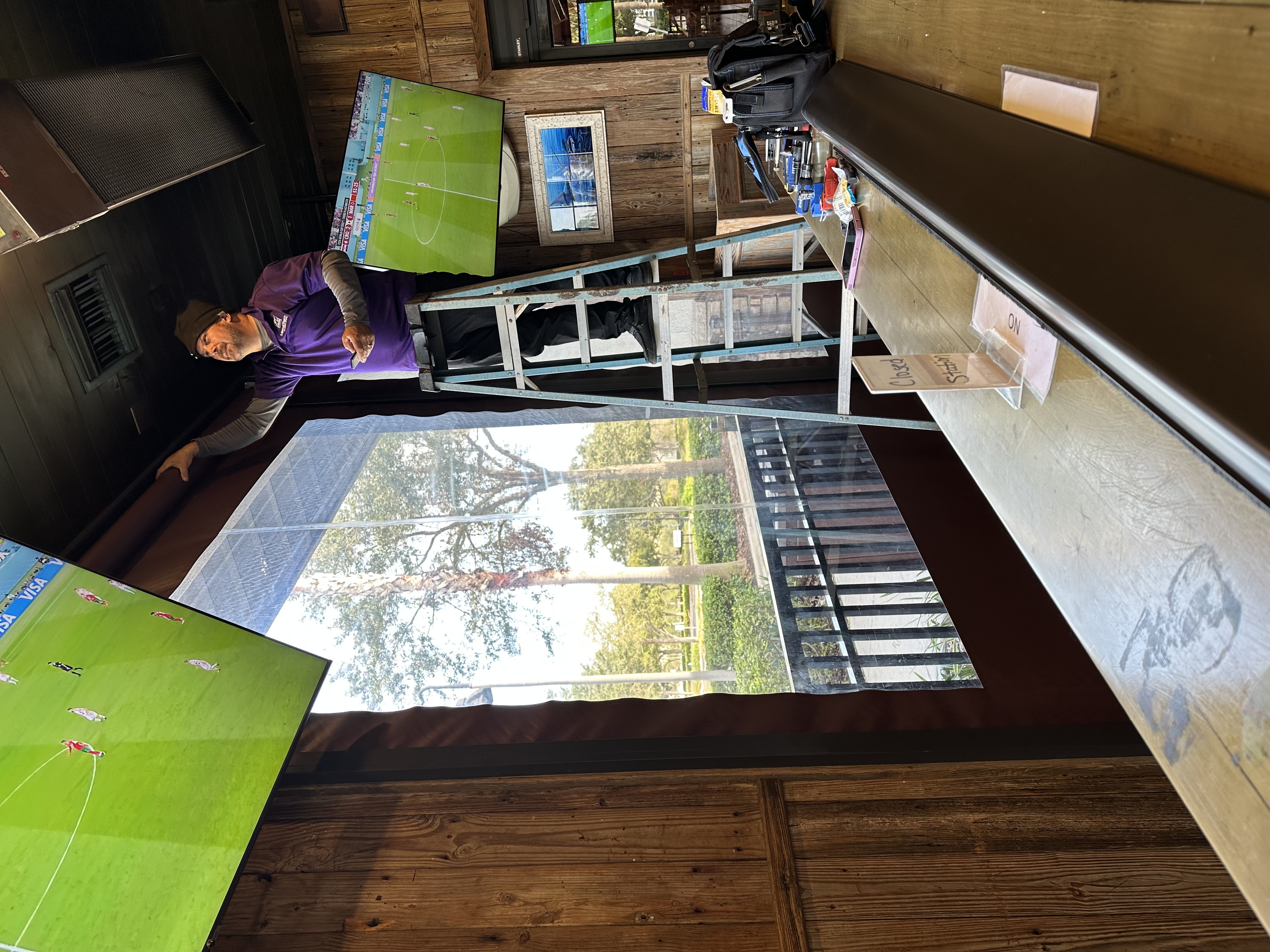 A Florida Living Outdoor Employee on a step ladder adjusts settings on a wall-mounted Motorized/Retractable screen in a bar with wood paneling and views of trees outside, enhanced by retractable screens for an unobstructed view.