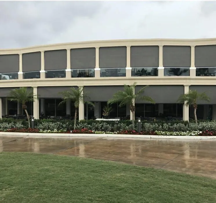 A two-story modern building featuring Solar shade retractable screens and surrounded by well-maintained landscaping, including palm trees and colorful flower beds under a cloudy sky, in a commercial environment. 