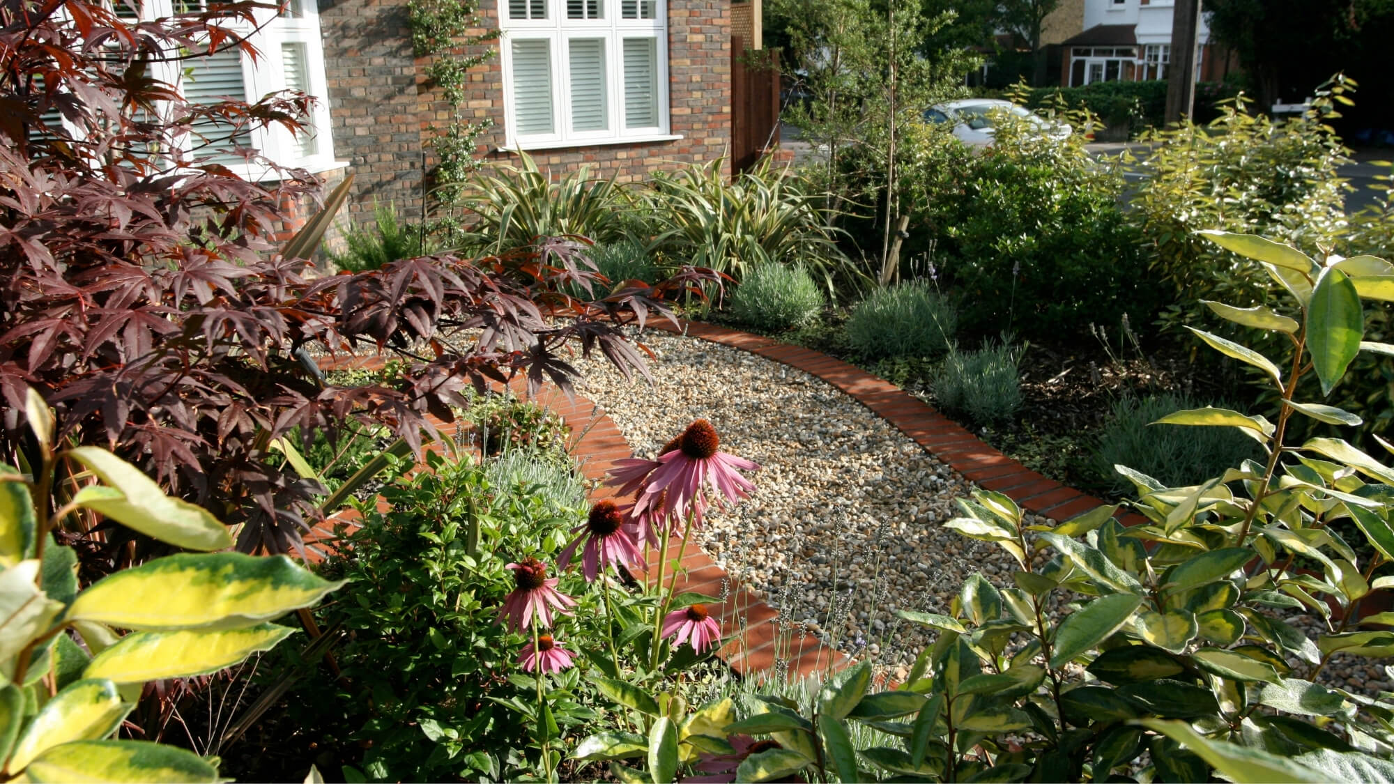 Low-maintenance front garden planting with curved natural gravel path and brick edging