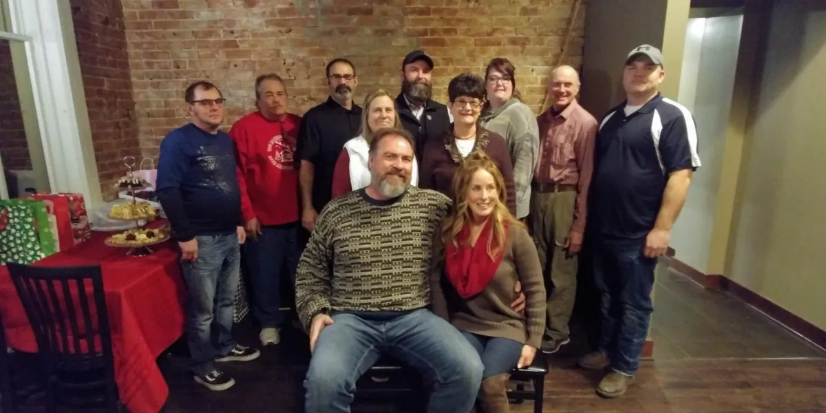 Group of eleven adults posing indoors at a holiday gathering with gifts and desserts on a table nearby.