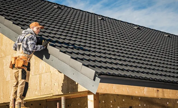 Roofer working on installing dark asphalt shingles on a residential roof under clear blue sky.