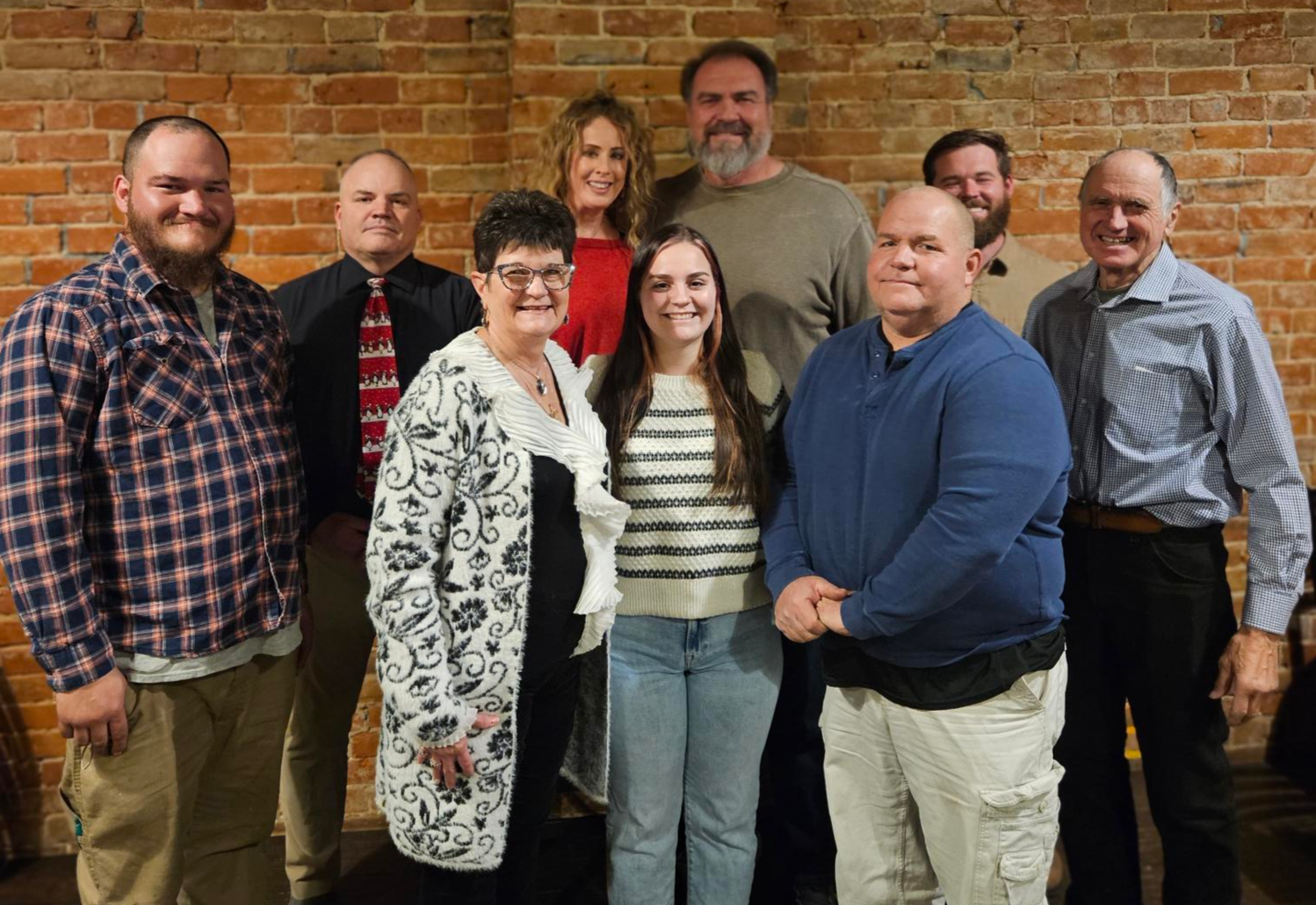Group of nine adults posing indoors at a holiday gathering with gifts and desserts on a table nearby.