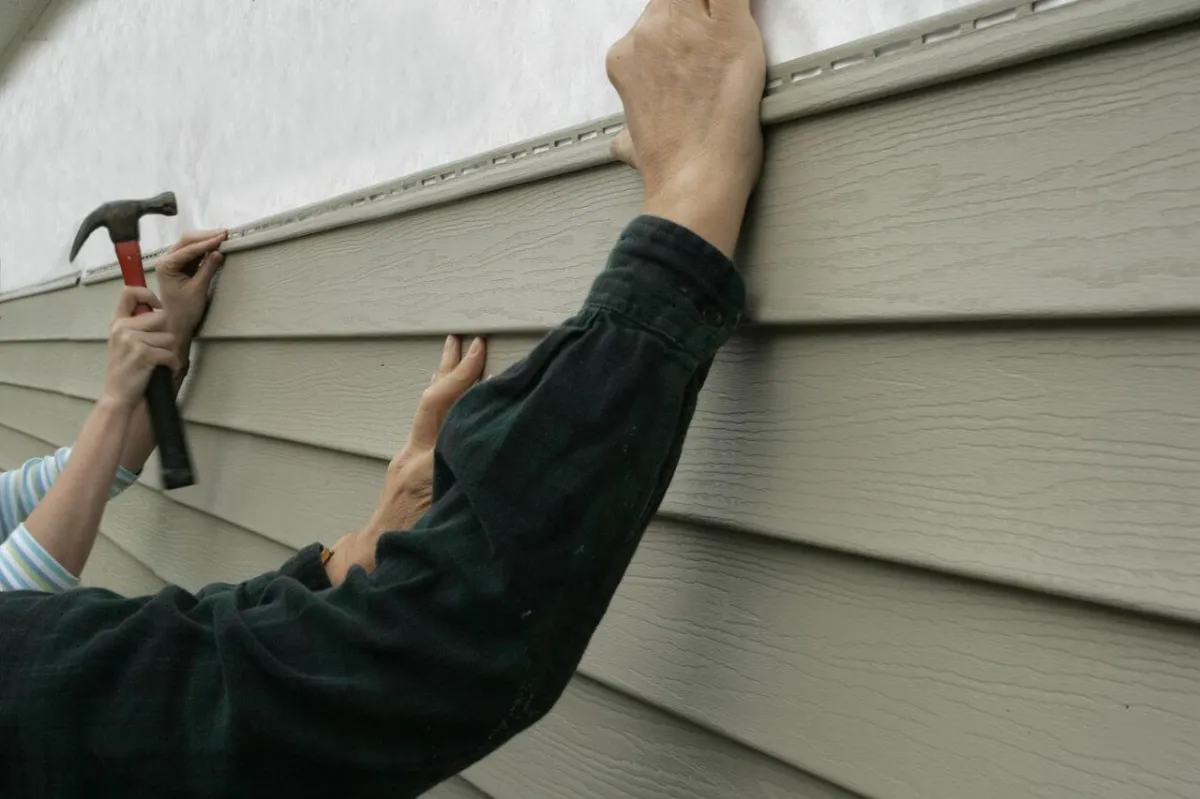 Hands installing gray vinyl siding on a house wall.