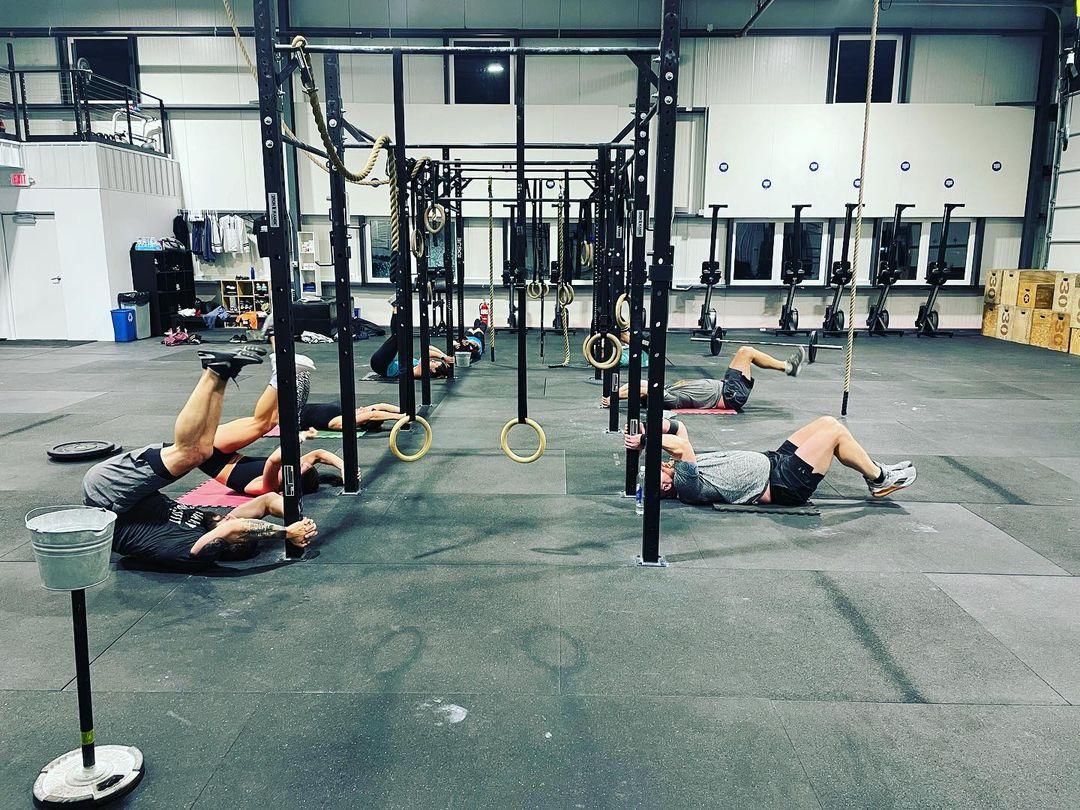 A diverse group of people participating in a high-energy CrossFit group class in Shelby. Participants are lifting weights, performing kettlebell swings, and doing box jumps in a well-equipped gym. The atmosphere is motivational, with everyone supporting each other during the workout