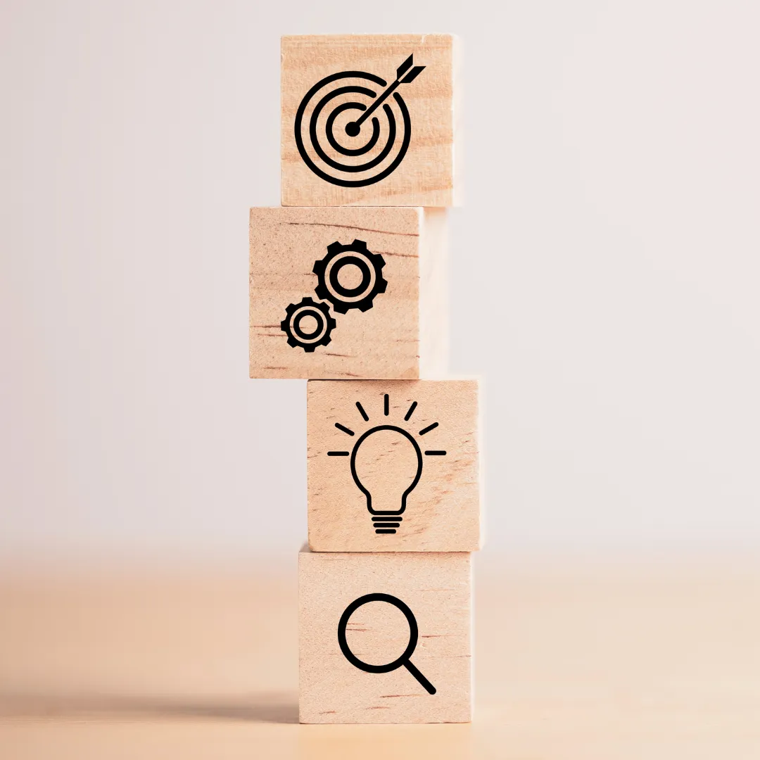 Stacked wooden blocks with icons of a magnifying glass, lightbulb, gears, and bullseye target, symbolizing strategy and goal setting.