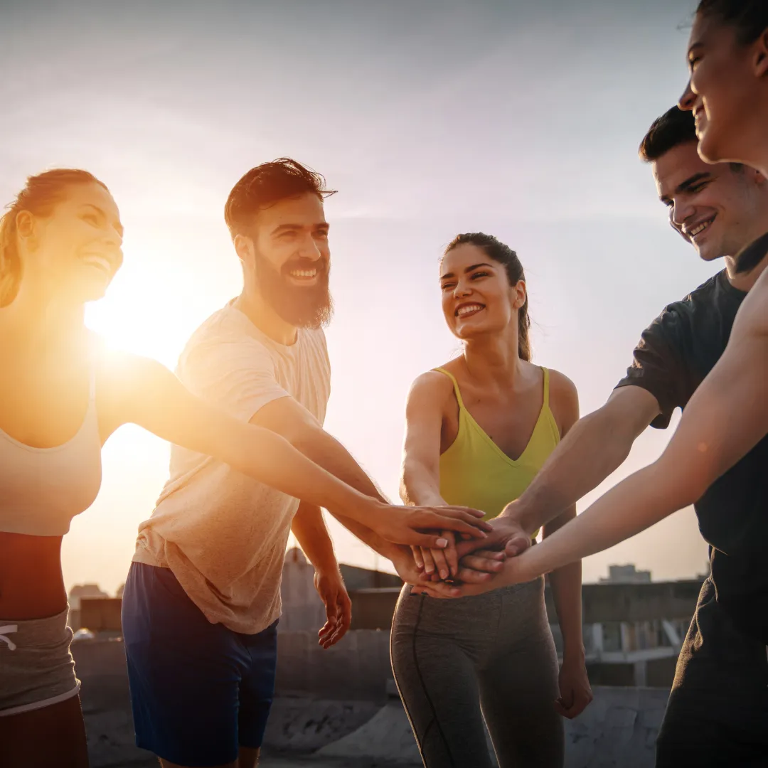 Group of young adults smiling and stacking hands together outdoors at sunset, showing teamwork and positivity.