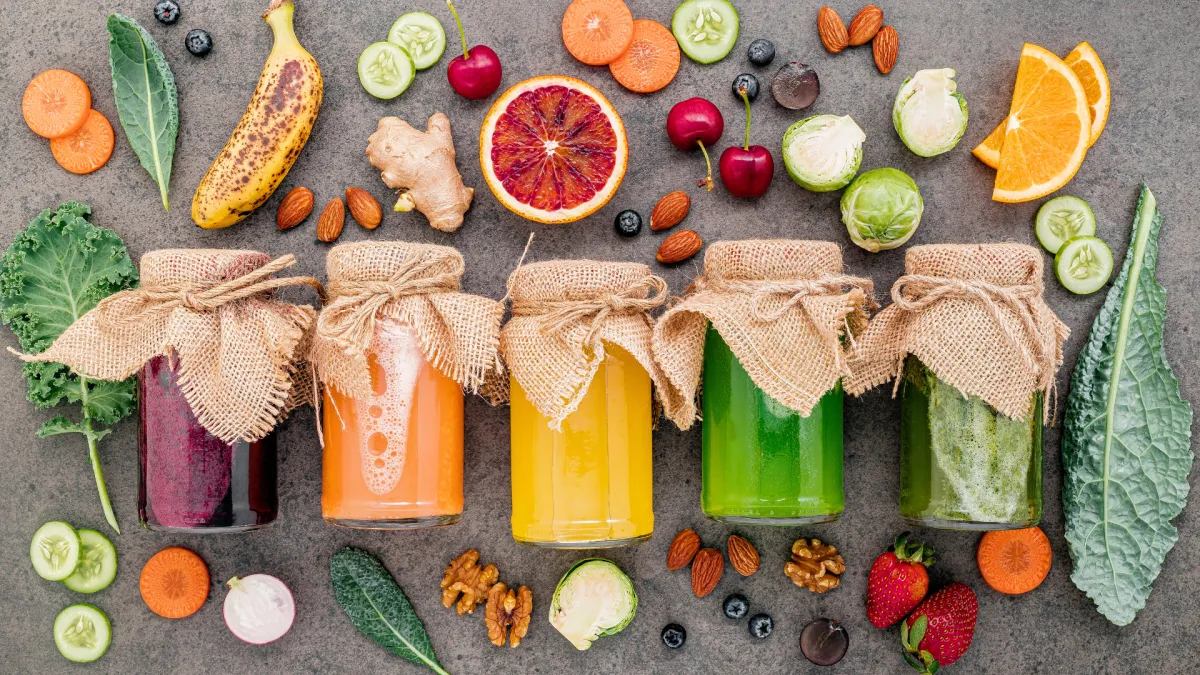 Colorful jars of juice surrounded by fresh fruits, vegetables, and nuts on a dark countertop.