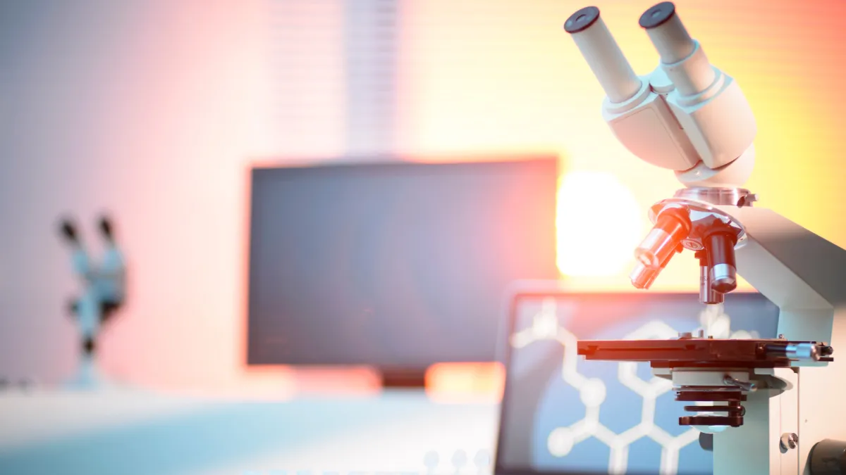 Microscope on a lab bench with test tubes and computer screens in a brightly lit modern laboratory.