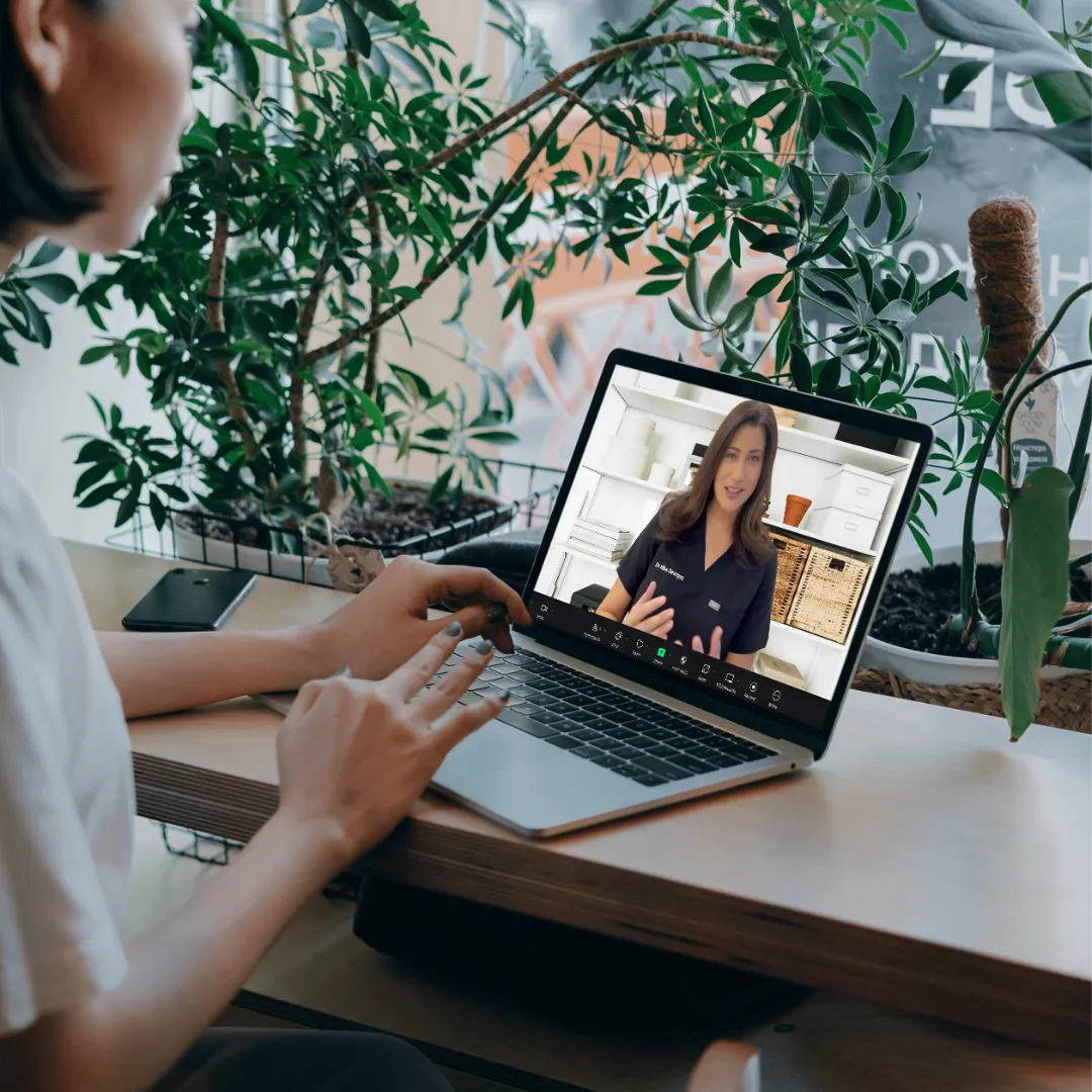 Woman in a video call with a doctor on a laptop, seated at a wooden desk surrounded by lush green plants.