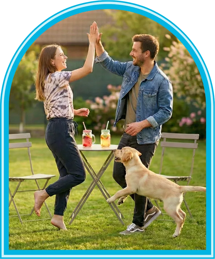 A couple enjoying a clean, pet-friendly backyard after a Clean Scoop dog waste removal service, high-fiving as their playful Labrador puppy jumps beside them.